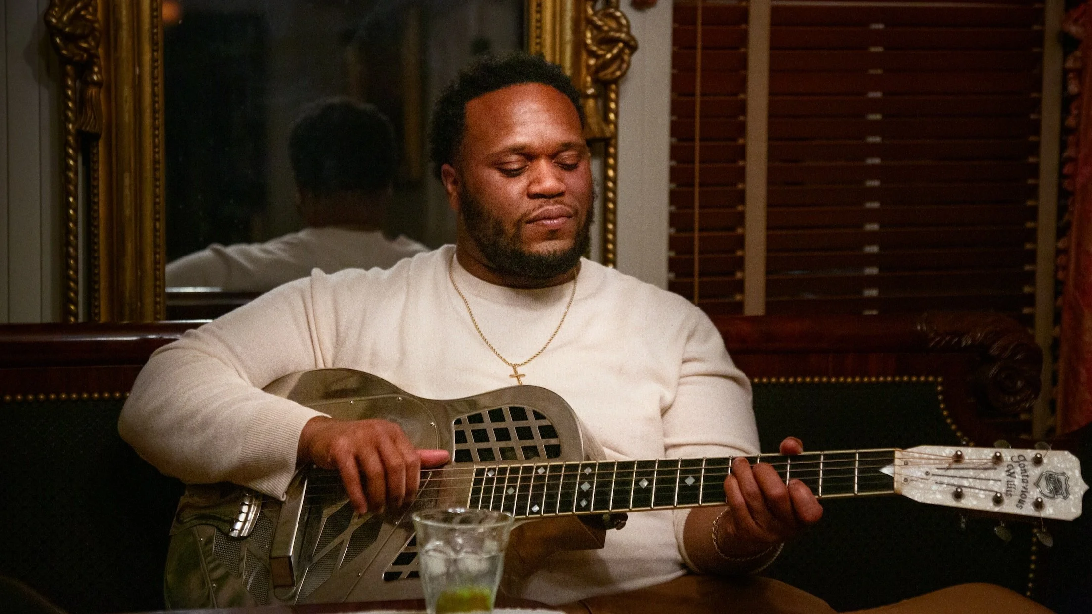 Man in a light sweater plays a resonator steel guitar indoors, wearing a gold cross necklace.